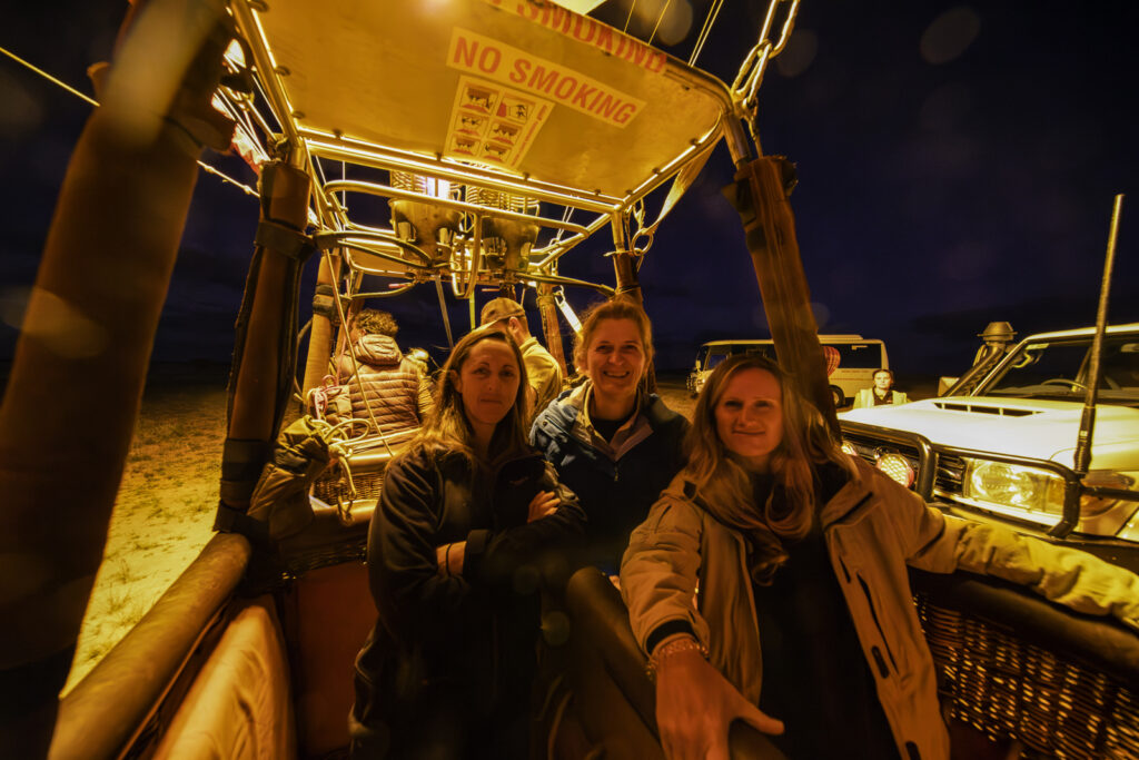 Experience the thrill of hot air ballooning with a happy trio in the stunning Outback near Alice Springs. Wrapped in warm coats and jackets, these adults enjoy a radiant morning, captured beautifully in this portrait. Feel the excitement of Outback Ballooning and remember to respect the no smoking rule for everyone's safety. Discover more at www.outbackballooning.com.au.